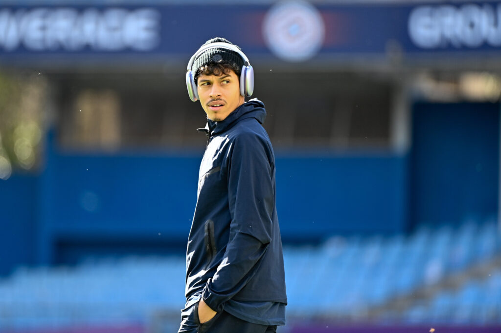 Young man wearing large over-ear headphones and a navy jacket stands on a blurred blue stadium backdrop, looking to his left.