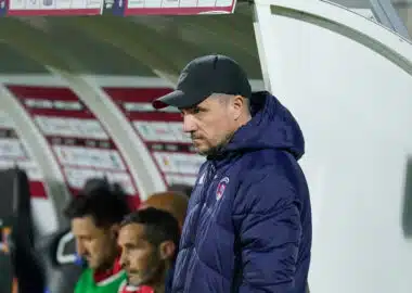 Side view of a man in a black cap and navy jacket standing near a dugout during a soccer match, with players seated behind him