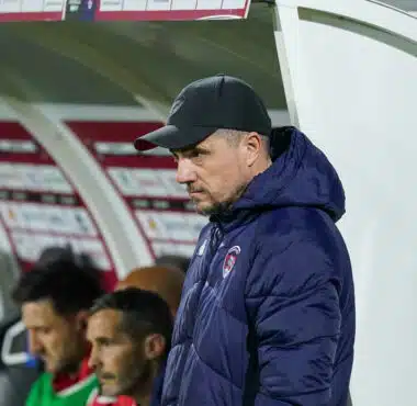Side view of a man in a black cap and navy jacket standing near a dugout during a soccer match, with players seated behind him