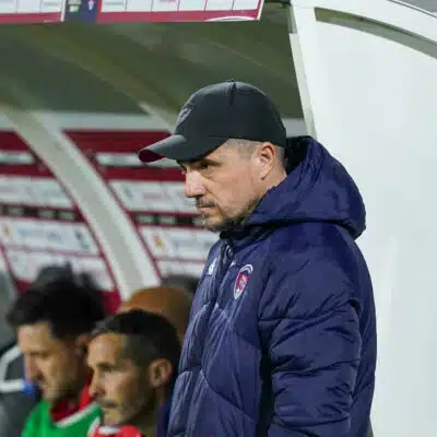 Side view of a man in a black cap and navy jacket standing near a dugout during a soccer match, with players seated behind him