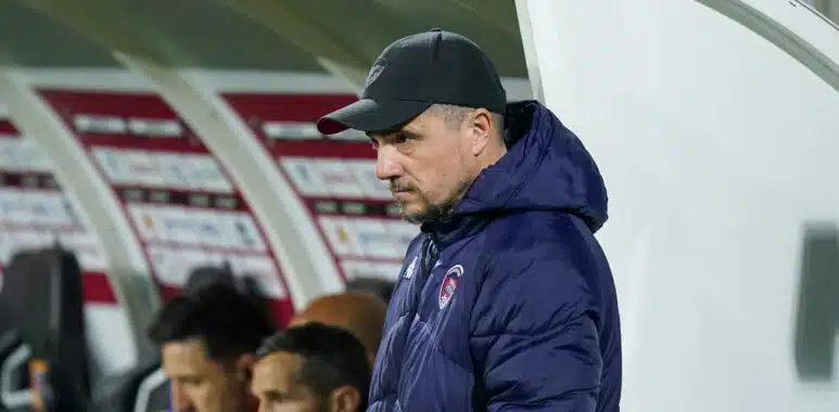 Side view of a man in a black cap and navy jacket standing near a dugout during a soccer match, with players seated behind him