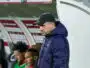 Side view of a man in a black cap and navy jacket standing near a dugout during a soccer match, with players seated behind him