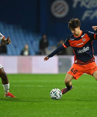 Two soccer players chase the ball on a green pitch during a match: one in white kit, the other in orange and navy.