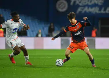 Two soccer players chase the ball on a green pitch during a match: one in white kit, the other in orange and navy.