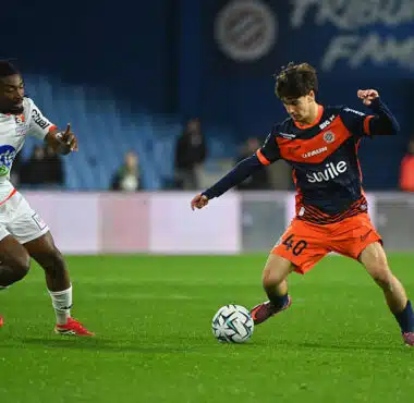 Two soccer players chase the ball on a green pitch during a match: one in white kit, the other in orange and navy.
