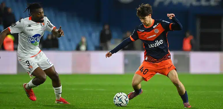 Vidal-Cartoux | AllezPaillade.com Two soccer players chase the ball on a green pitch during a match: one in white kit, the other in orange and navy.