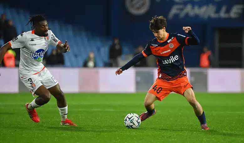 Two soccer players chase the ball on a green pitch during a match: one in white kit, the other in orange and navy.