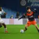 Two soccer players chase the ball on a green pitch during a match: one in white kit, the other in orange and navy.