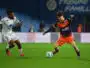 Two soccer players chase the ball on a green pitch during a match: one in white kit, the other in orange and navy.