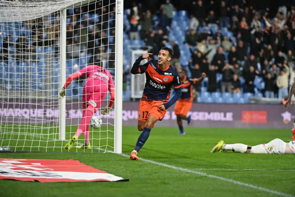 Soccer player in navy and orange celebrates near the goal after scoring, with a pink-clad goalkeeper on the ground and a cheering crowd in the stands.