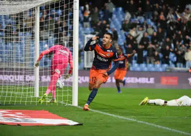 Soccer player in navy and orange celebrates near the goal after scoring, with a pink-clad goalkeeper on the ground and a cheering crowd in the stands.