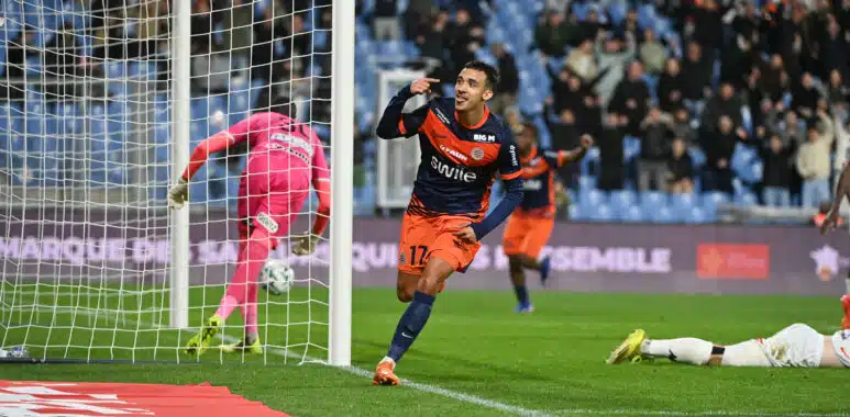 Soccer player in navy and orange celebrates near the goal after scoring, with a pink-clad goalkeeper on the ground and a cheering crowd in the stands.