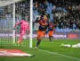 Soccer player in navy and orange celebrates near the goal after scoring, with a pink-clad goalkeeper on the ground and a cheering crowd in the stands.