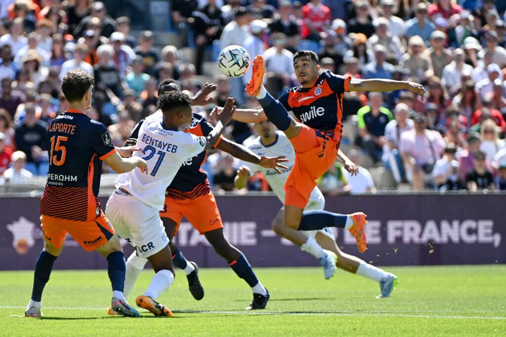 Soccer players in orange and dark uniforms contest a ball in midair during a crowded match on a sunny day.