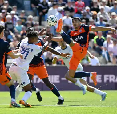 Soccer players in orange and dark uniforms contest a ball in midair during a crowded match on a sunny day.