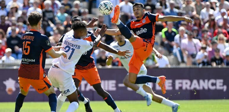 Soccer players in orange and dark uniforms contest a ball in midair during a crowded match on a sunny day.