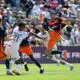 Soccer players in orange and dark uniforms contest a ball in midair during a crowded match on a sunny day.
