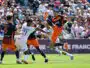 Soccer players in orange and dark uniforms contest a ball in midair during a crowded match on a sunny day.