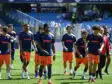 Soccer players in blue jerseys and orange shorts walk onto the field before a game, PROMAN logo visible on shirts and a stadium in the background.