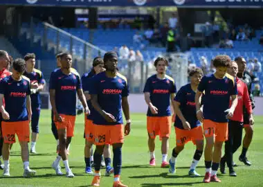 Soccer players in blue jerseys and orange shorts walk onto the field before a game, PROMAN logo visible on shirts and a stadium in the background.