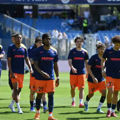Soccer players in blue jerseys and orange shorts walk onto the field before a game, PROMAN logo visible on shirts and a stadium in the background.
