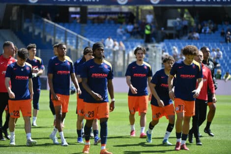 Soccer players in blue jerseys and orange shorts walk onto the field before a game, PROMAN logo visible on shirts and a stadium in the background.