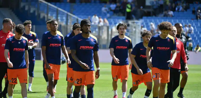 Soccer players in blue jerseys and orange shorts walk onto the field before a game, PROMAN logo visible on shirts and a stadium in the background.