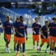Soccer players in blue jerseys and orange shorts walk onto the field before a game, PROMAN logo visible on shirts and a stadium in the background.