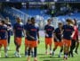 Soccer players in blue jerseys and orange shorts walk onto the field before a game, PROMAN logo visible on shirts and a stadium in the background.