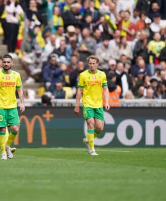 Soccer players in yellow and green kits walk on a grass field as a crowded stadium watches the match.