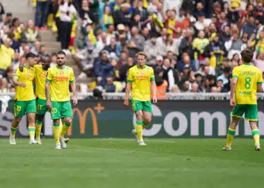 Soccer players in yellow and green kits walk on a grass field as a crowded stadium watches the match.