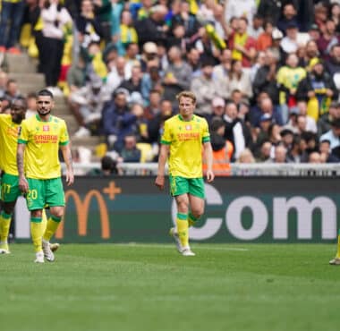 Soccer players in yellow and green kits walk on a grass field as a crowded stadium watches the match.