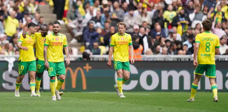Soccer players in yellow and green kits walk on a grass field as a crowded stadium watches the match.