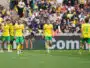 Soccer players in yellow and green kits walk on a grass field as a crowded stadium watches the match.