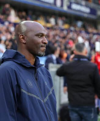 Man in a navy zip-up jacket stands at the sideline of a football match, with spectators in the stands behind him.