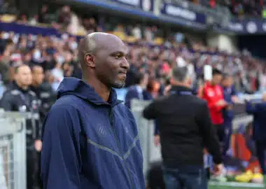 Man in a navy zip-up jacket stands at the sideline of a football match, with spectators in the stands behind him.