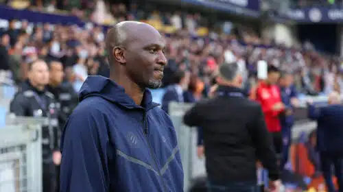 Man in a navy zip-up jacket stands at the sideline of a football match, with spectators in the stands behind him.
