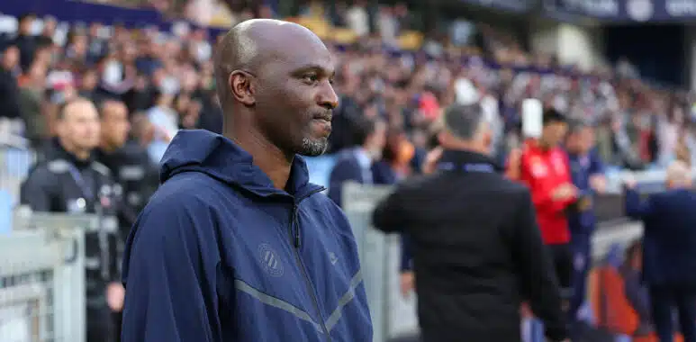 Man in a navy zip-up jacket stands at the sideline of a football match, with spectators in the stands behind him.