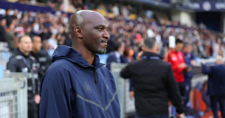 Man in a navy zip-up jacket stands at the sideline of a football match, with spectators in the stands behind him.