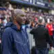 Man in a navy zip-up jacket stands at the sideline of a football match, with spectators in the stands behind him.