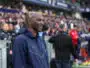 Man in a navy zip-up jacket stands at the sideline of a football match, with spectators in the stands behind him.