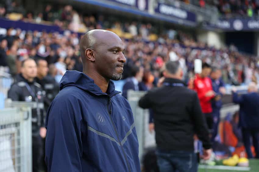 Zoumana Camara | AllezPaillade.com Man in a navy zip-up jacket stands at the sideline of a football match, with spectators in the stands behind him.