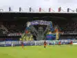 Giant mural of three hooded figures holding a banner above players on a soccer pitch in a stadium.
