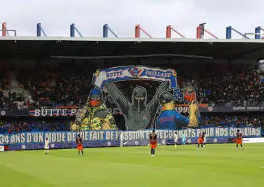 Butte Paillade Ultras fumigène | AllezPaillade.com Giant mural of three hooded figures holding a banner above players on a soccer pitch in a stadium.