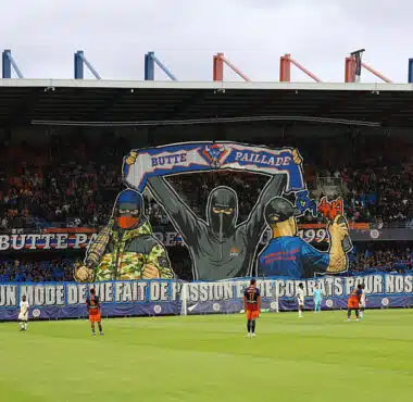 Giant mural of three hooded figures holding a banner above players on a soccer pitch in a stadium.