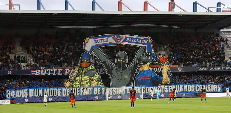 Giant mural of three hooded figures holding a banner above players on a soccer pitch in a stadium.