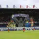 Giant mural of three hooded figures holding a banner above players on a soccer pitch in a stadium.