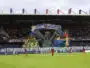 Giant mural of three hooded figures holding a banner above players on a soccer pitch in a stadium.