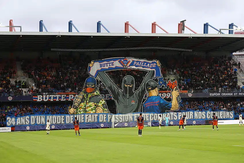 Giant mural of three hooded figures holding a banner above players on a soccer pitch in a stadium.