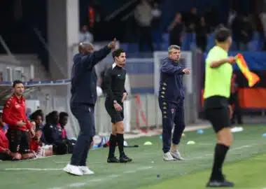Football sideline scene: coach in black with headset stands while another coach in a navy tracksuit points, substitutes in red sit on the bench.